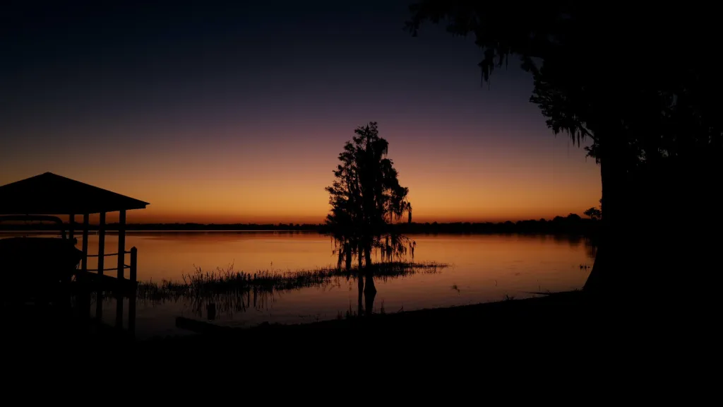 Sunset silhouette over lake with dock pavilion and cypress tree, orange and purple sky